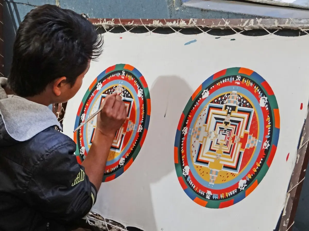 A young man with a fine brush painting a round mandala of red, blue, green and gold rings and squares on a white canvas, beside a finished mandala of the same design