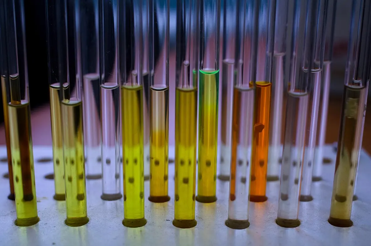 A row of thin glass tubes standing upright in a white rack, filled to different heights with clear, yellow, green and orange liquids