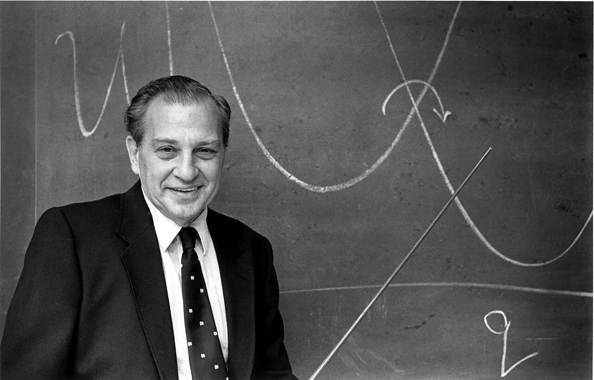A smiling man in a dark suit and tie stands at a blackboard holding a long pointer. Behind him are chalk drawings of two U-shaped curves that cross, with a curved arrow near the crossing, and a horizontal line below.