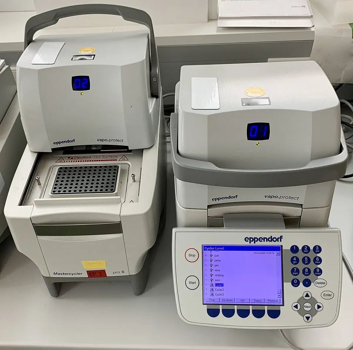Two gray laboratory machines on a bench. The lid of one is lifted, showing a metal block full of small holes for tubes; the other has a small blue screen and a keypad on the front