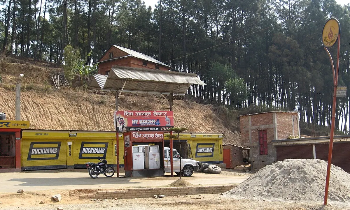 A small roadside petrol station with two white fuel pumps under a metal roof, a parked white car and a motorbike, yellow walls with Duckhams signs, and pine trees on the hill behind