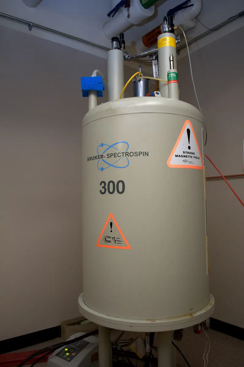 A tall cream-colored cylinder taller than a person, on a round stand in a small room, with pipes on top and orange warning stickers saying strong magnetic field