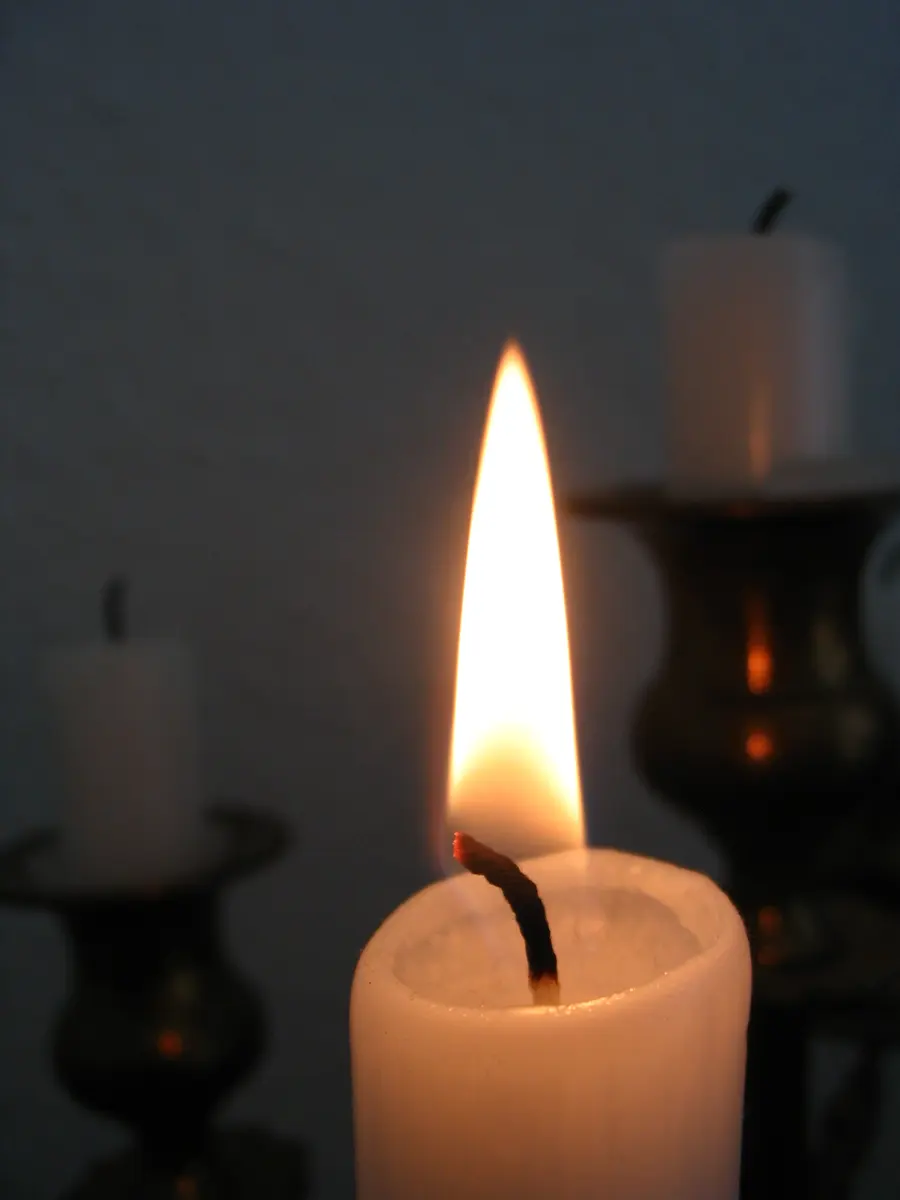 A burning white candle in the front, its wick bent, with two unlit white candles in dark candlesticks behind it