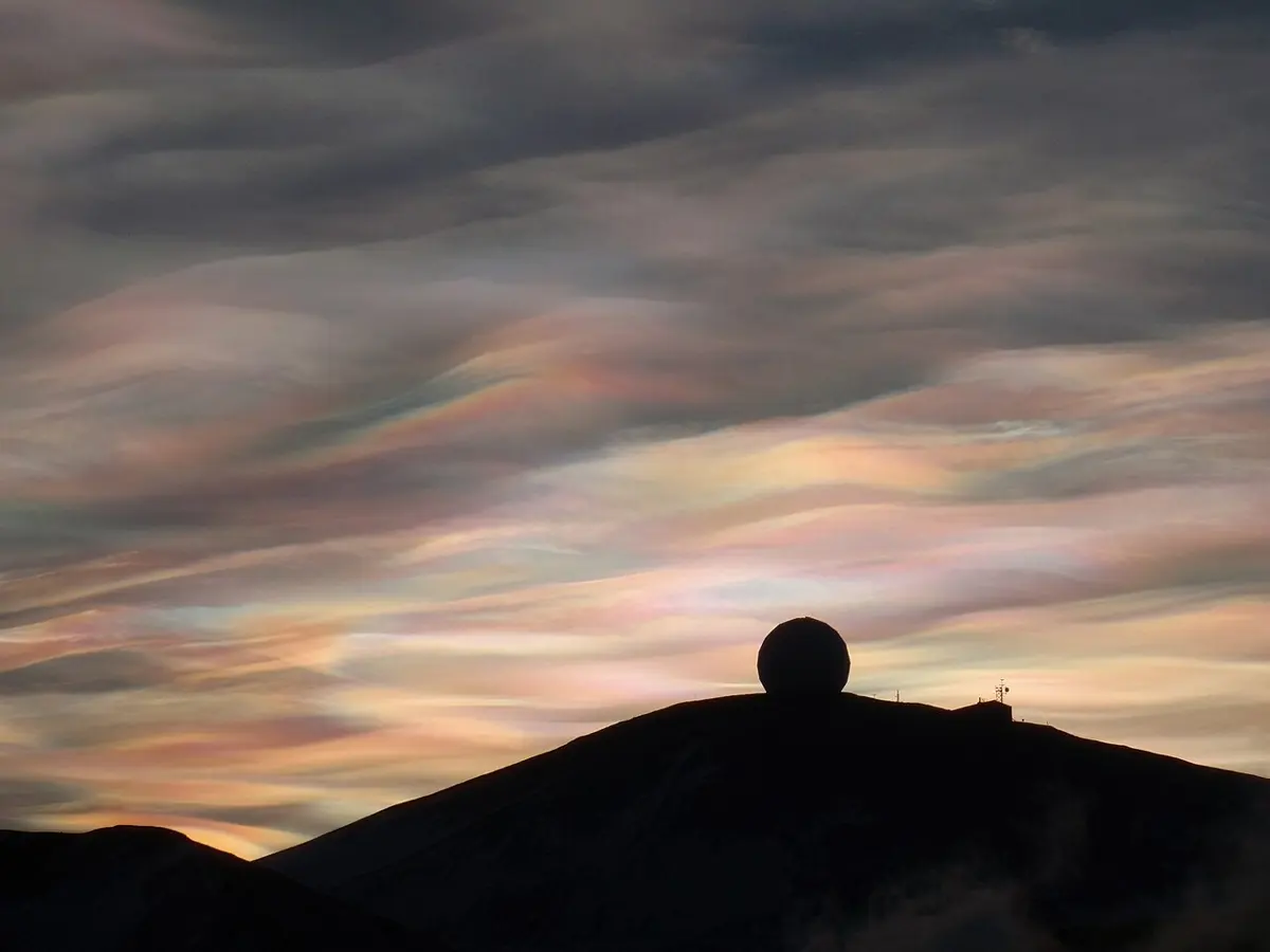 Wavy clouds shimmering in soft pink, green and gold over a dark hill topped by a white dome, against a gray sky