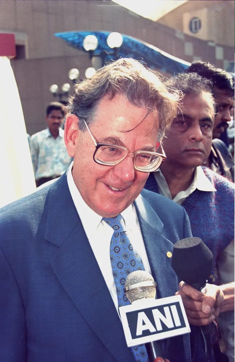 A smiling gray-haired man in glasses, a suit and a blue tie, surrounded by people holding microphones towards him, outdoors in a crowd