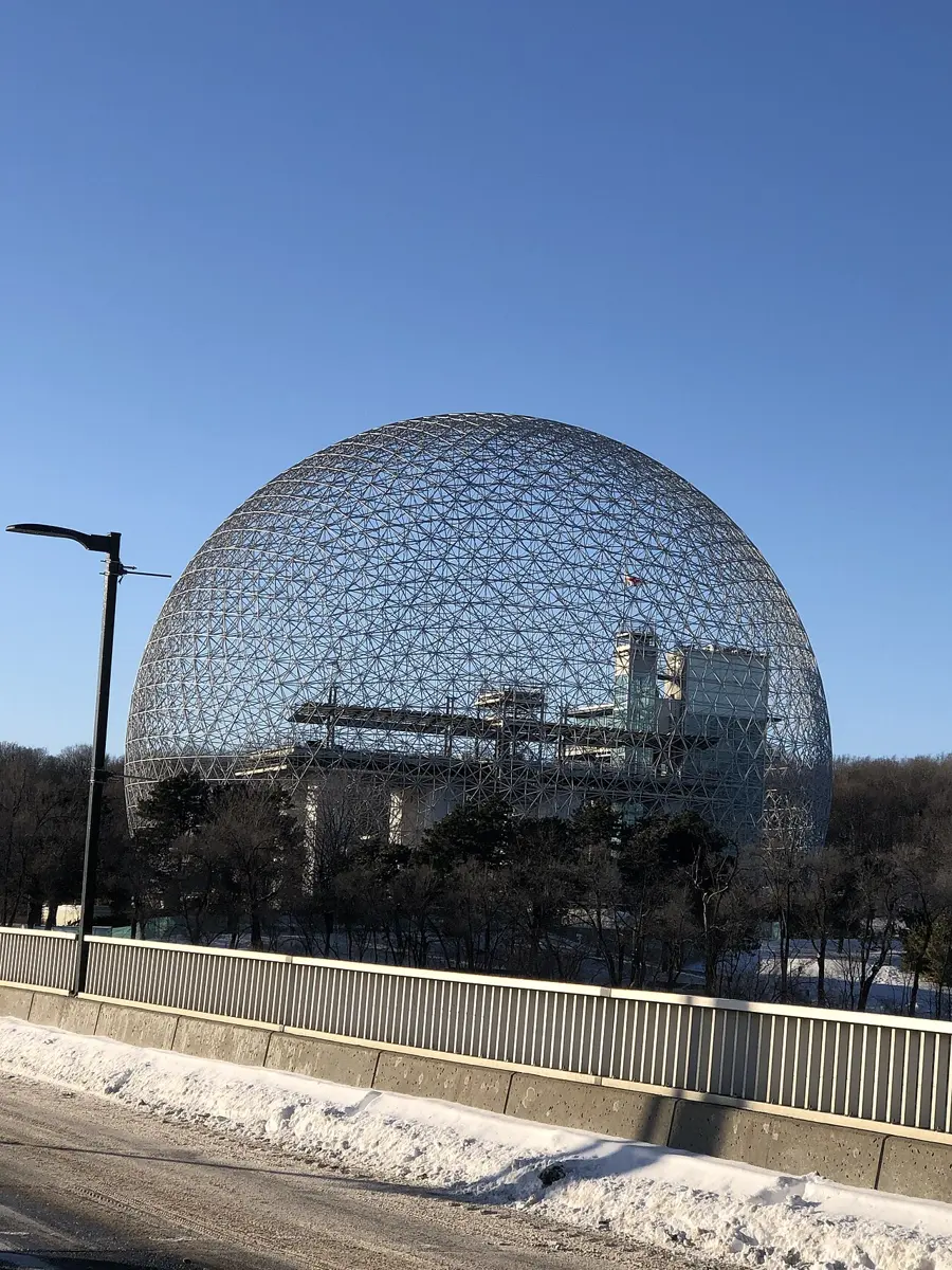 A huge, almost round dome made of a lattice of thin metal struts, with buildings visible inside it, standing beyond bare trees and a snowy road under a clear blue sky