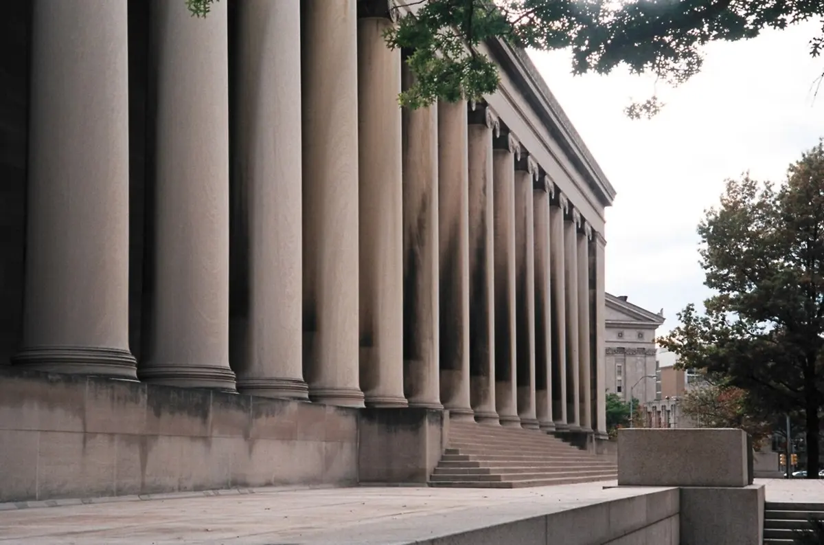 A long row of tall stone columns along the side of a large gray building, with steps and trees