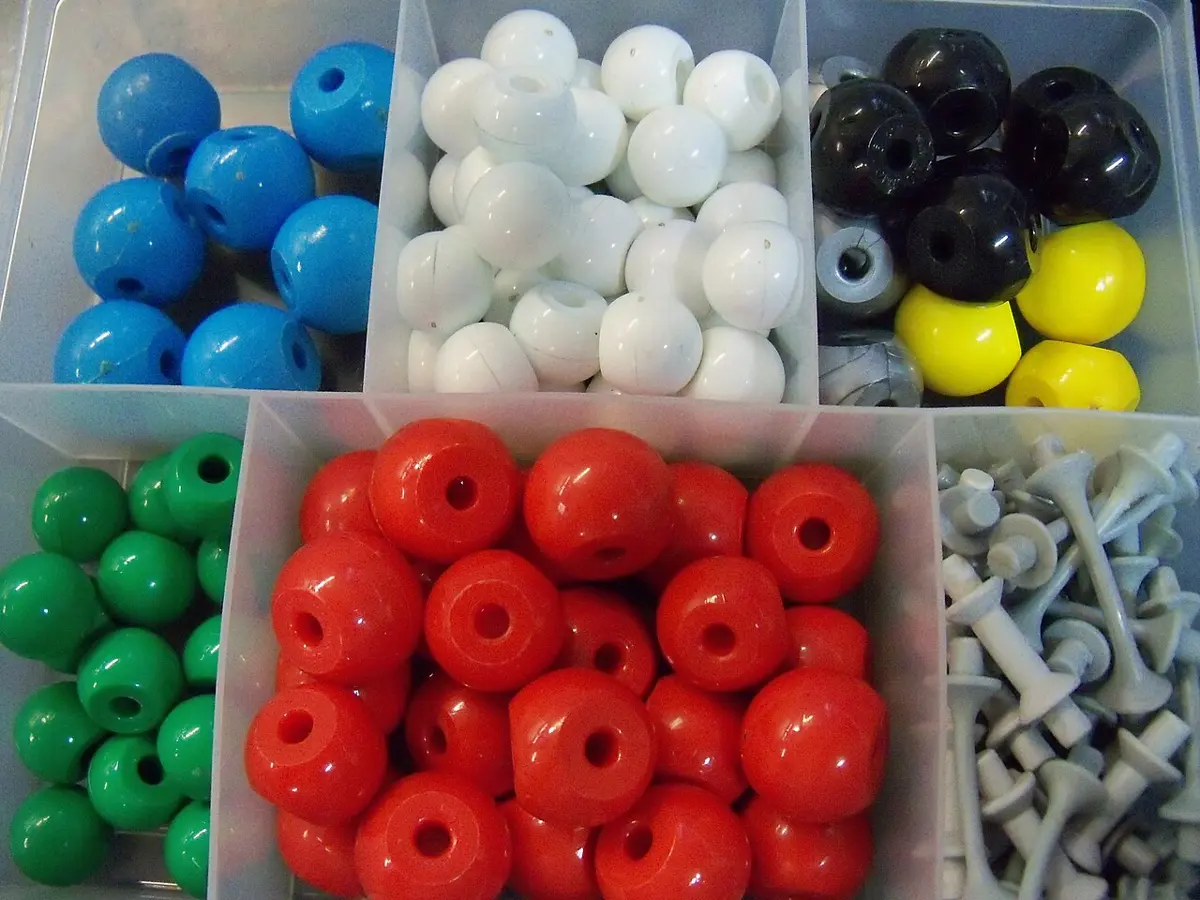 A plastic box divided into compartments full of colored plastic balls with holes in them, blue, white, black, yellow, green and red, and a compartment of small gray connecting pieces