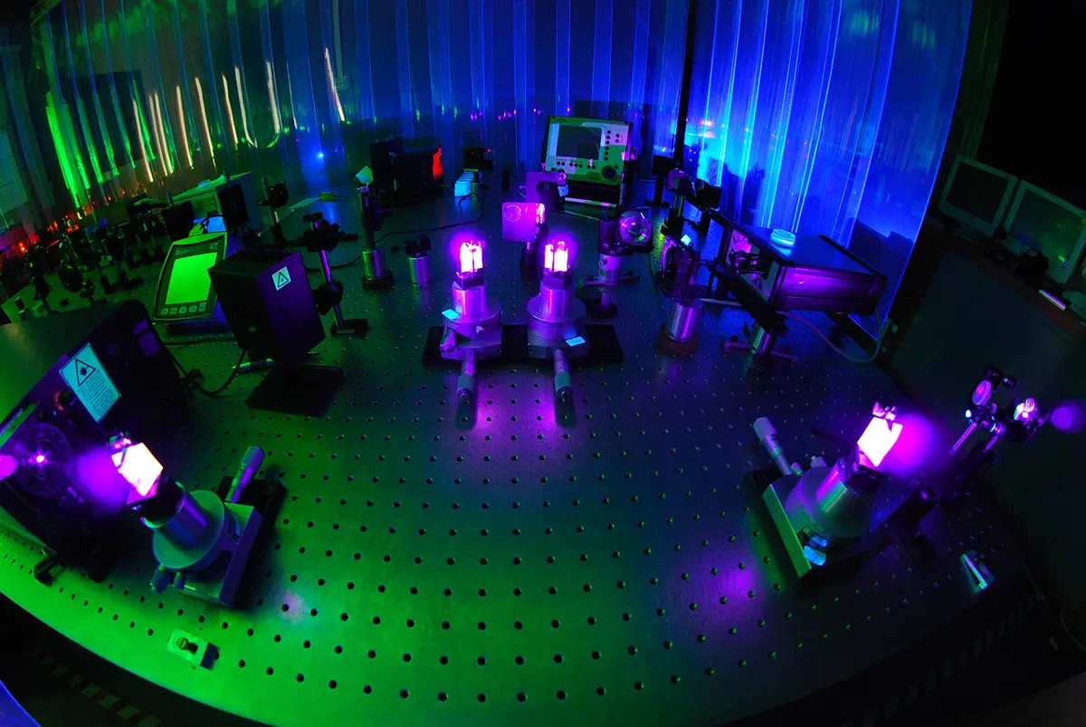 A dark laboratory with a large metal table full of holes, on which stand mirrors and crystals on posts, some glowing violet in the laser light