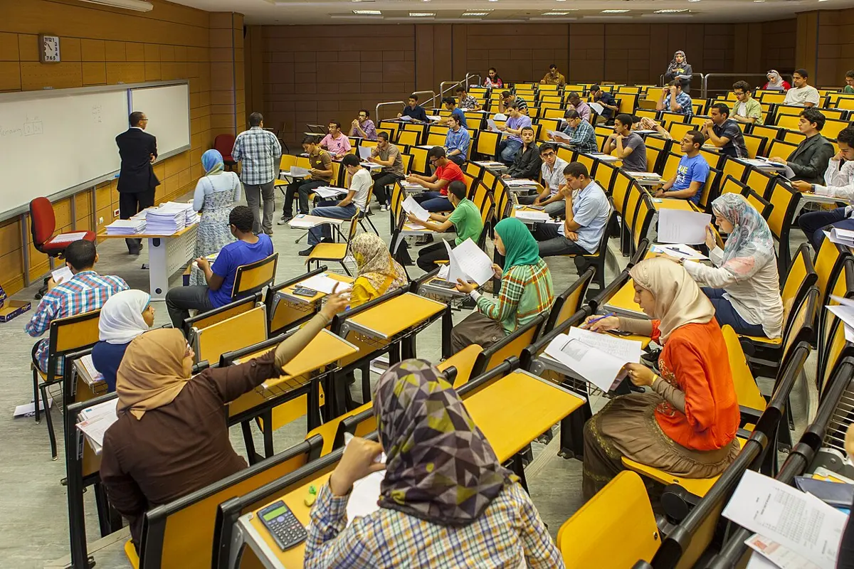 Students at desks in rows of yellow seats in a large lecture hall, working on papers, with a teacher at the whiteboard