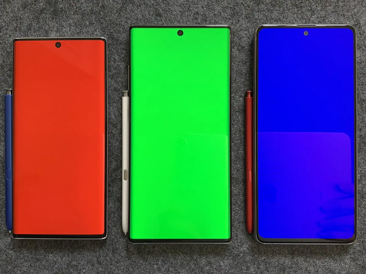 Three smartphones lying on a gray cloth, each with a stylus beside it, their screens glowing plain red, plain green and plain blue