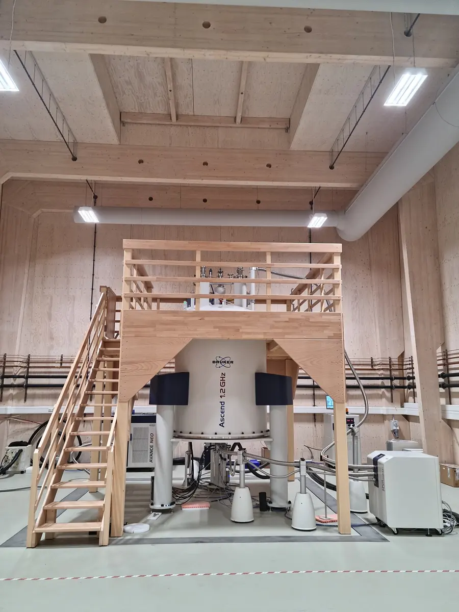 A huge white cylinder labeled Ascend 1.2 GHz, standing on legs in a tall wooden hall, with a wooden platform and staircase built around its top so people can reach it