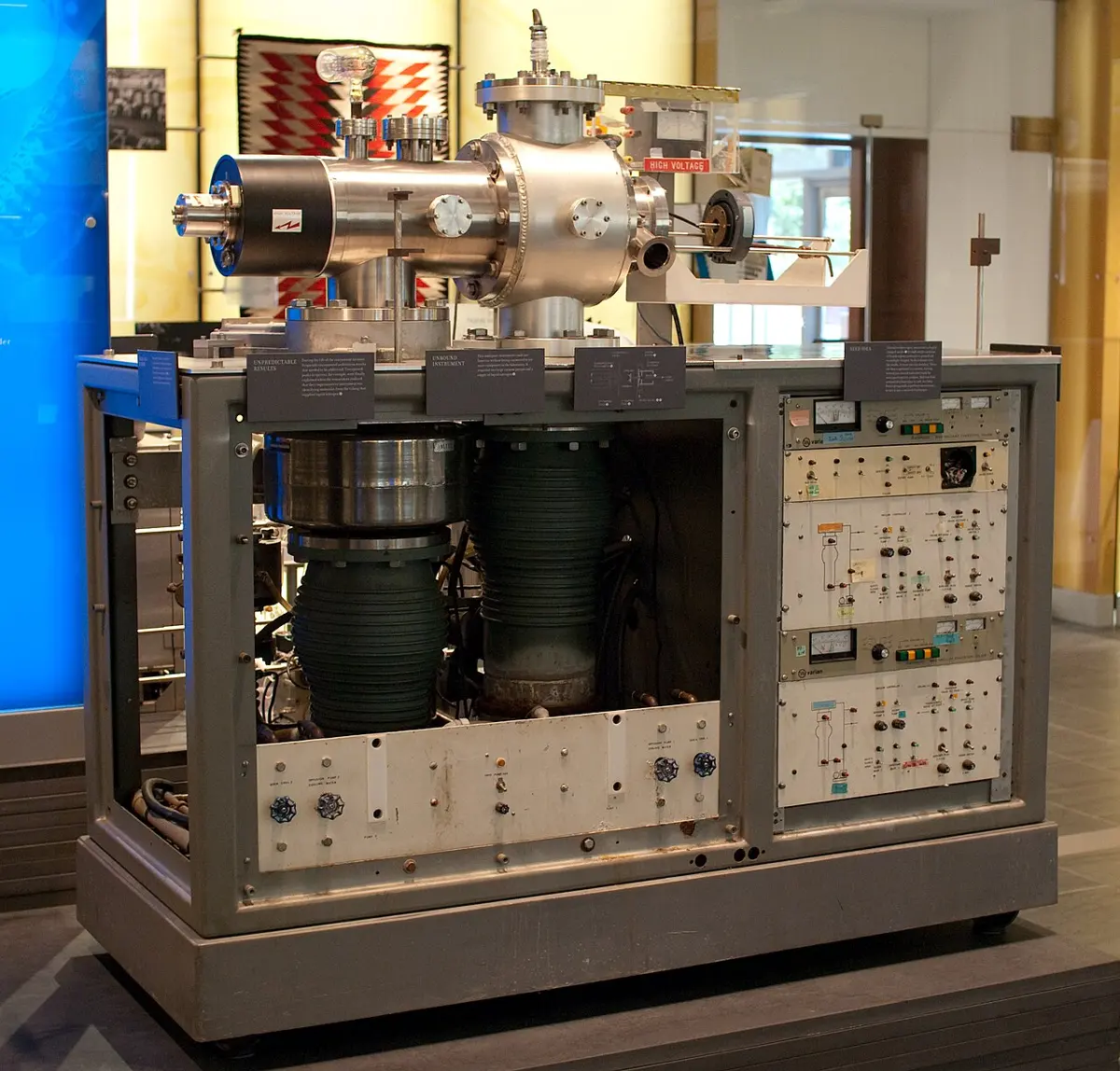 A tall gray cabinet in a museum display: panels of switches, dials and meters, with shiny steel vacuum chambers and pipes on top