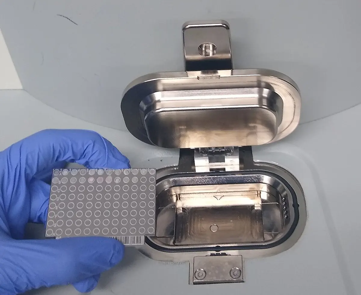 A hand in a blue glove holding a small steel plate marked with a grid of tiny round sample spots, next to the open metal holder that carries it into the machine
