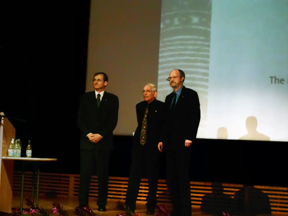 Three men in dark suits standing on a stage in front of a large screen: a younger man with dark hair on the left, an older white-haired man in the middle, and a bearded man on the right