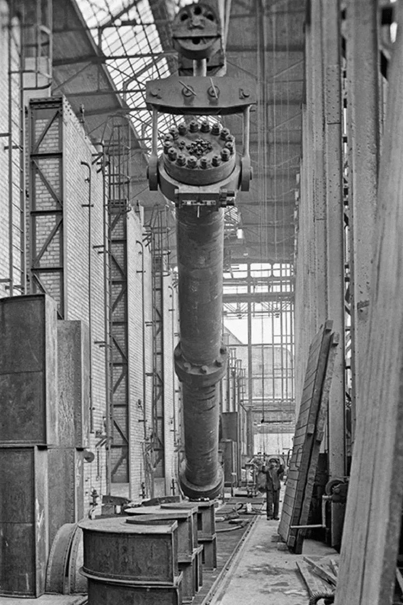 Black-and-white photo of a tall, thick steel tube hanging upright from a crane inside a factory hall, with a man standing at its foot