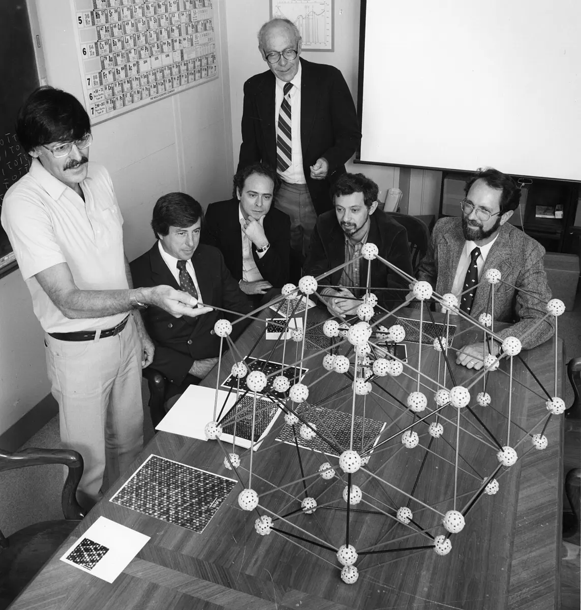 Black-and-white photo of six men around a table with a large ball-and-stick model of atoms; the man standing on the left points at the model