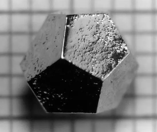 A small, shiny, silvery grain of metal shaped like a dodecahedron, with flat five-sided faces, lying on graph paper