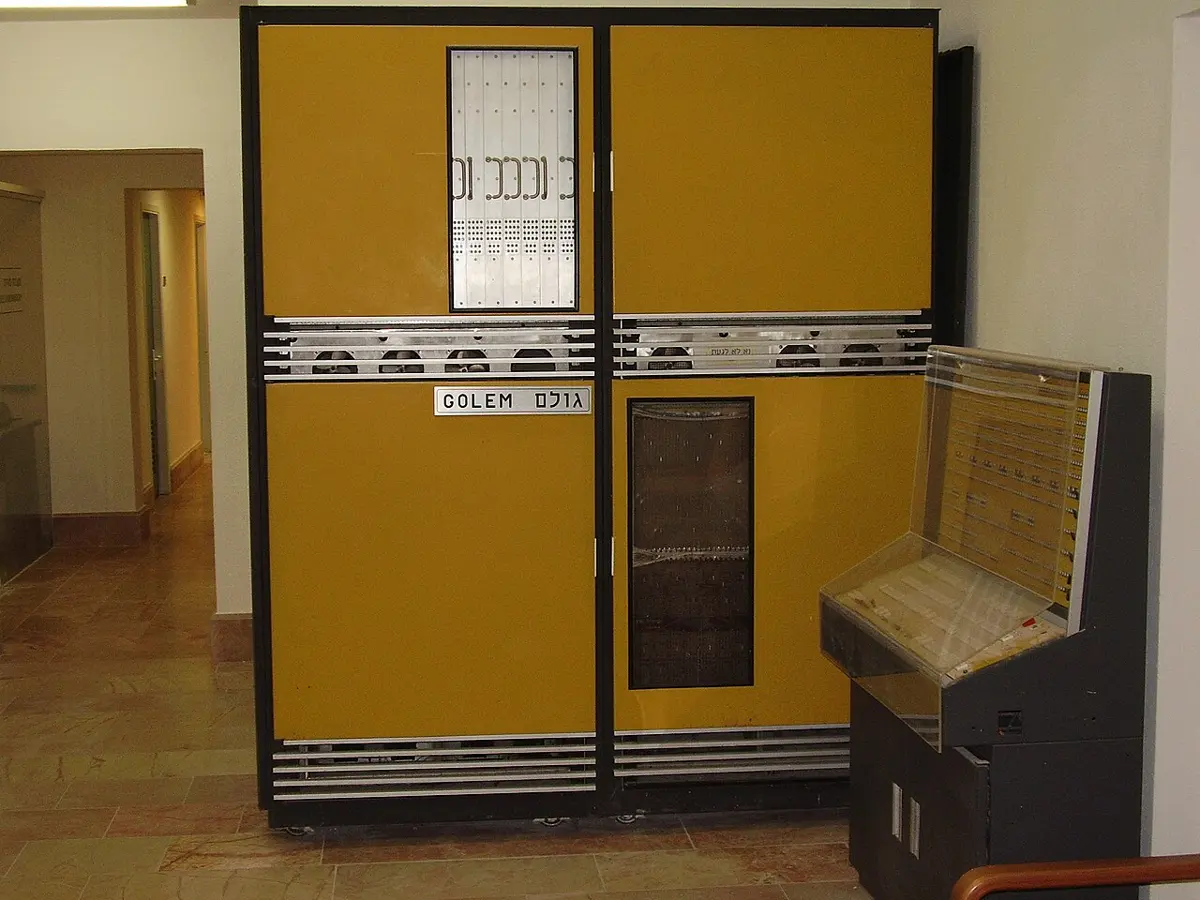 A mustard-yellow metal cabinet, taller than a person, with a sign reading GOLEM in English and Hebrew letters, standing in a corridor beside a sloping console with rows of switches