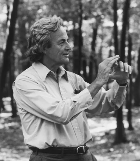 Black-and-white photo of a gray-haired man in a light shirt, standing in a wood and holding up his hands as if shaping something small