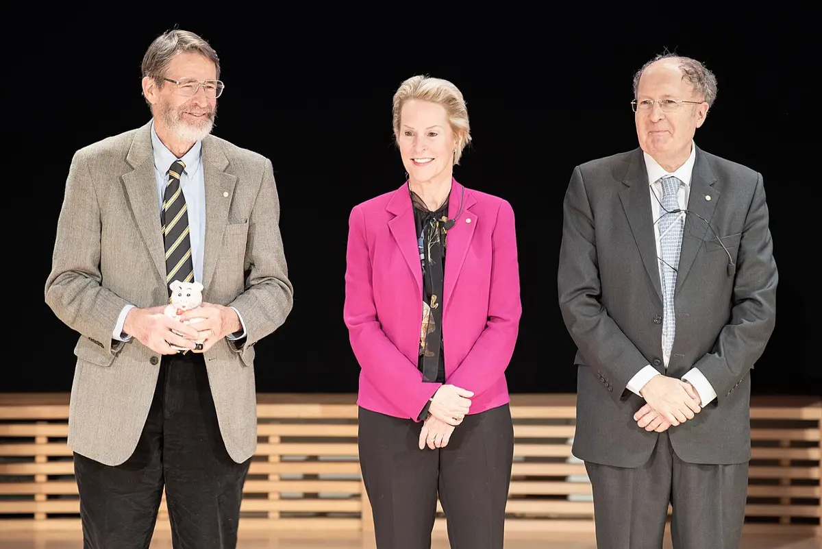 Three people standing side by side on a stage and smiling: a man with a white beard in a tweed jacket, a woman in a pink jacket, and a man in a gray suit