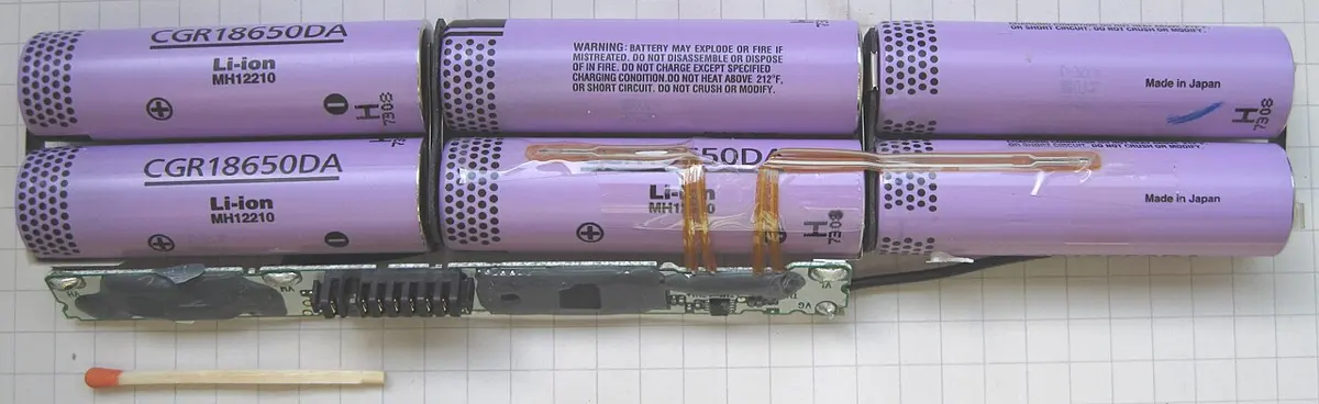 An opened laptop battery: six purple cylindrical cells in two rows, joined by metal strips, with a long thin circuit board below them and a matchstick beside it for scale