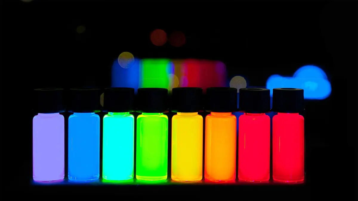 A row of eight small bottles glowing in the dark, from violet on the left through blue, green, yellow and orange to deep red on the right