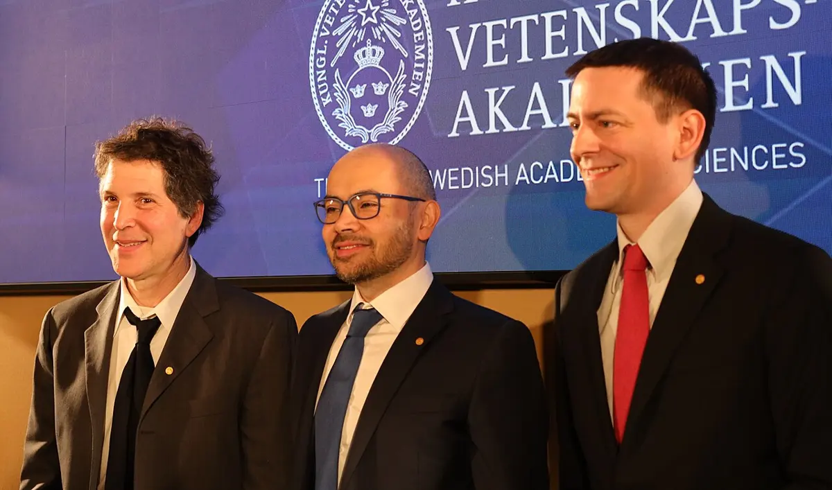 Three smiling men in dark suits standing side by side in front of a blue banner of the Royal Swedish Academy of Sciences