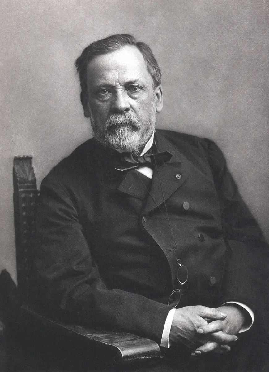 A black-and-white studio portrait of an older man with a gray beard and a bow tie, sitting in a carved chair with his hands folded, looking at the camera