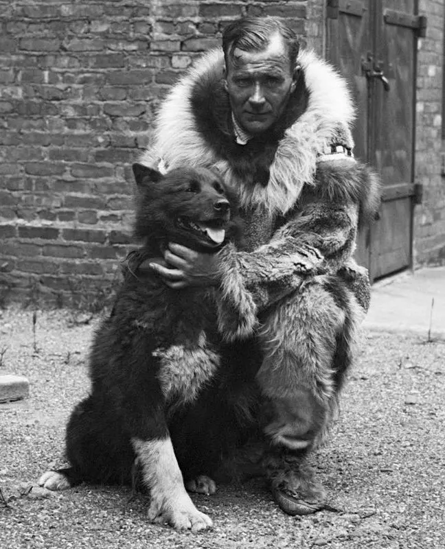 An old black-and-white photograph of a man in a thick fur parka crouching beside a dark, fluffy husky dog in front of a brick wall, his arm round the dog