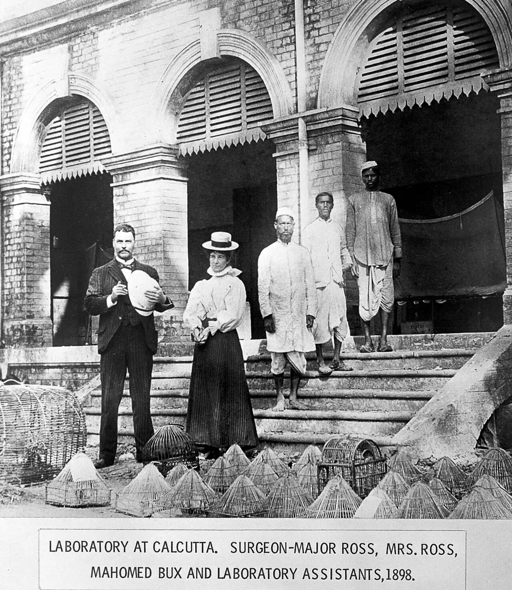 An old photograph of a man in a dark suit and a woman in a white blouse and hat standing on the steps of a brick building with arched windows, with three Indian men behind them, and small dome-shaped net cages on the ground in front