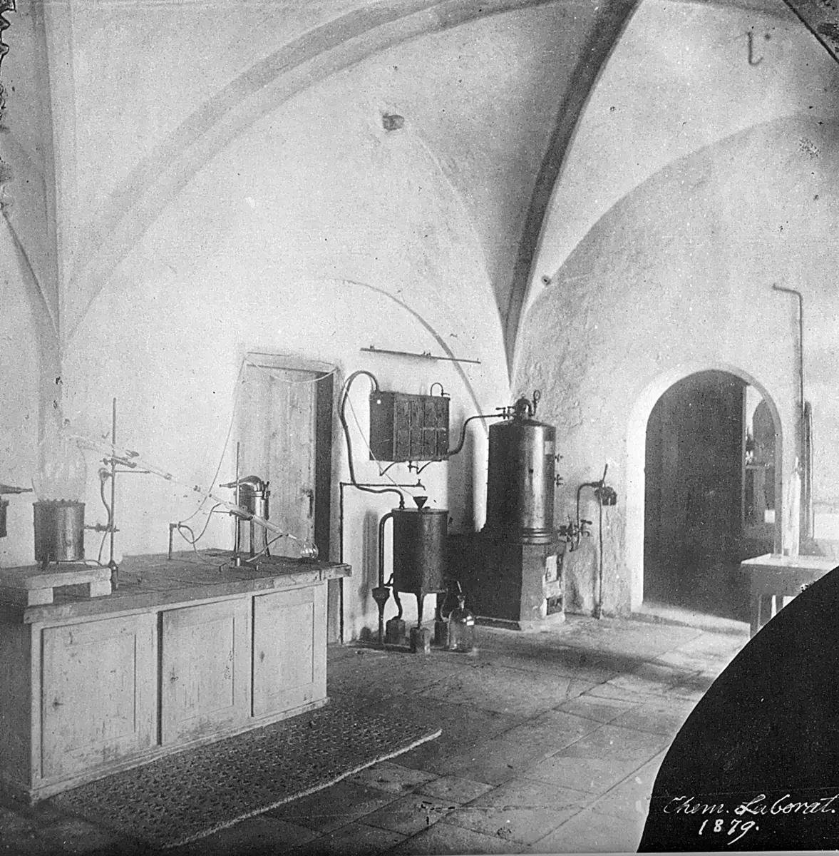 An old black-and-white photograph of a room with a high, curved stone ceiling, a wooden workbench with glass flasks and tubes on stands, and metal tanks and pipes along the white walls
