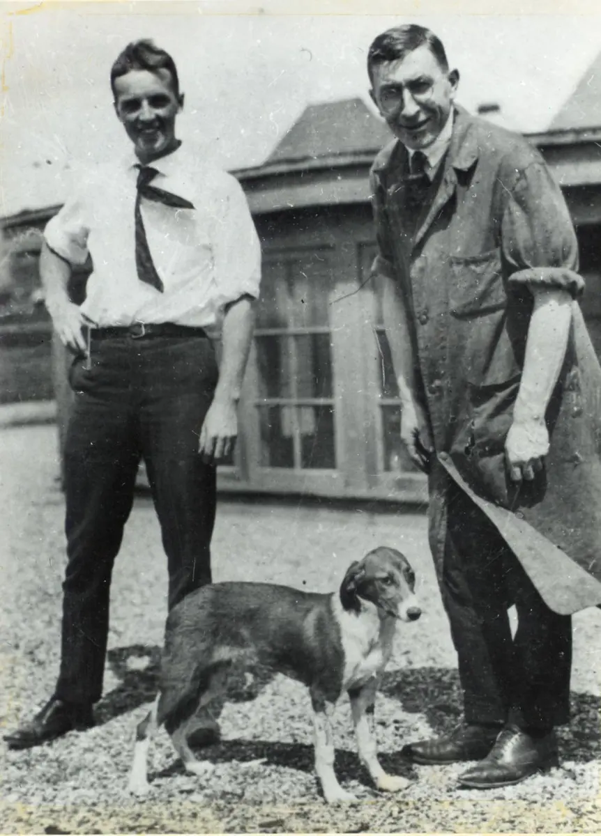 Two men standing on a flat roof beside a brick building, a young man in a white shirt and tie and a taller man in a long laboratory coat, with a brown and white dog standing in front of them