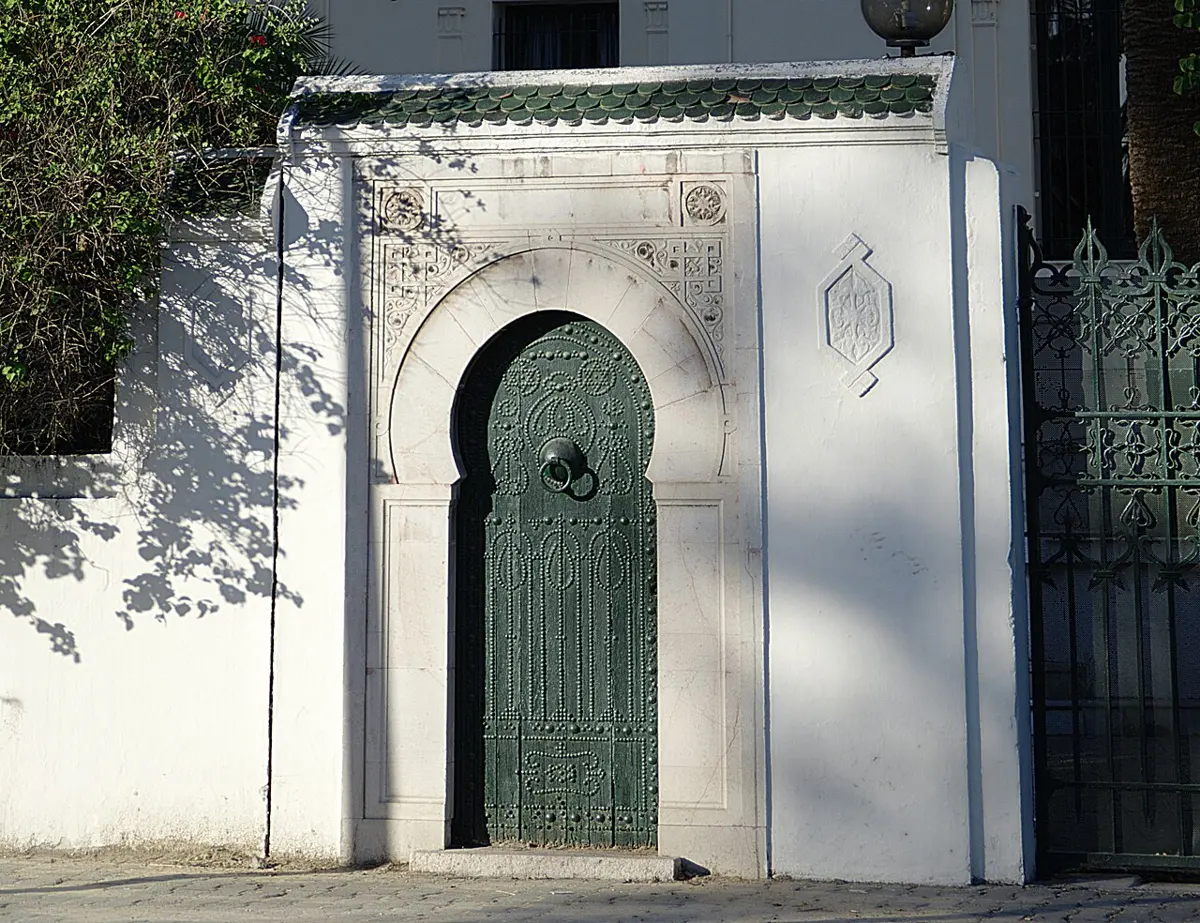 A white wall with a horseshoe-shaped arched doorway framed in carved stone, and a green carved wooden door, beside a green iron gate