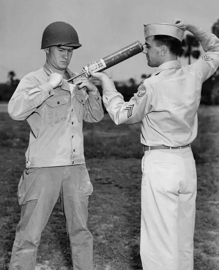 An old black-and-white photograph of two American soldiers in a field: one holds a long hand sprayer and puffs powder into the open front of the other soldier’s shirt