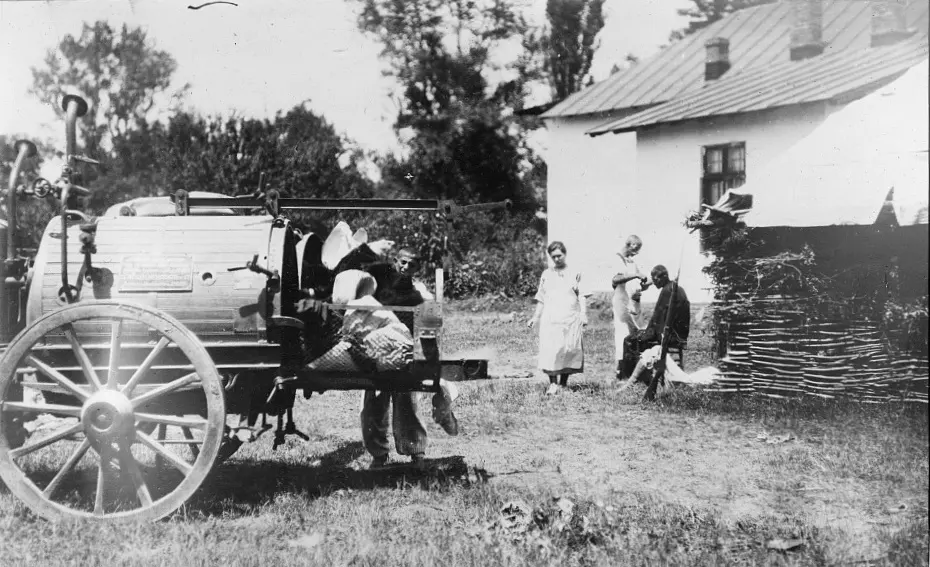 An old black-and-white photograph of a large round metal tank on big spoked wheels, standing in a grassy yard beside a white cottage, with a few people nearby