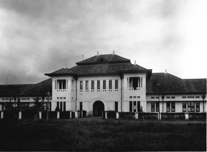 An old black-and-white photograph of a long, white two-storey building with a tiled roof and a tall central entrance arch, behind a lawn