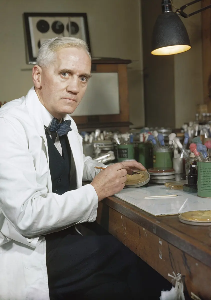 Alexander Fleming in a white coat and bow tie, sitting at his laboratory bench with culture dishes, holding one in his hands