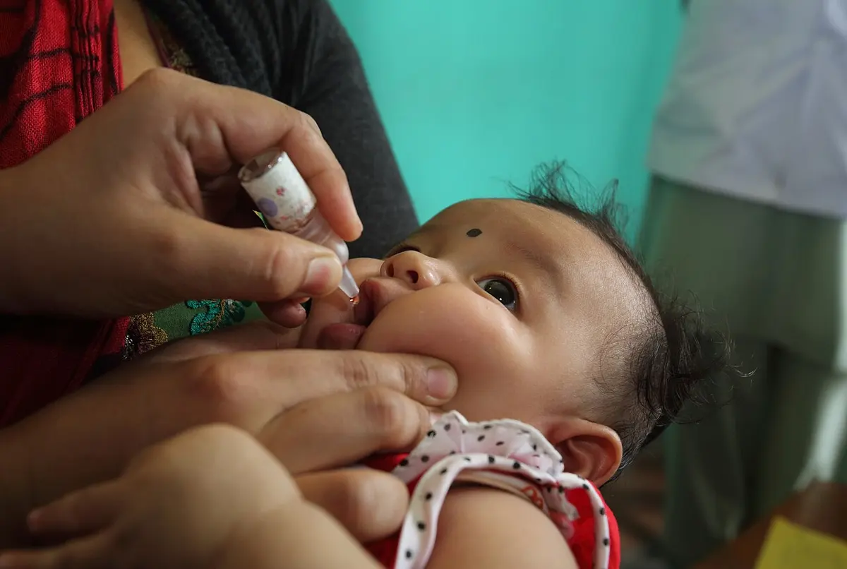 A close-up of a baby held in someone's arms, mouth open, as a hand squeezes a drop of vaccine from a small plastic dropper bottle into the baby's mouth.