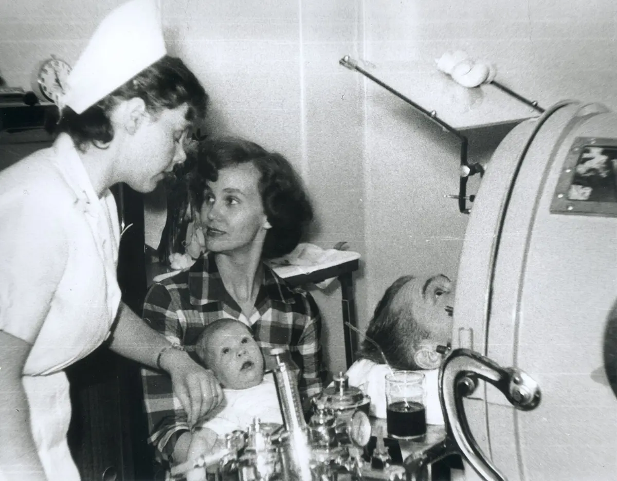 An old black and white photograph: a man lies inside a large metal cylinder with only his head outside, resting on a pillow; a nurse in a white cap and a woman holding a baby lean over to talk to him.
