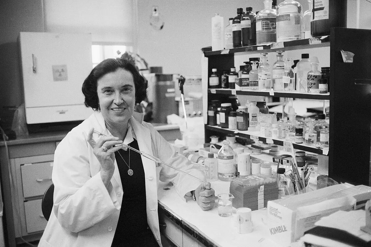 A smiling woman in a white lab coat sits at a laboratory bench, holding a long glass pipette, with shelves of bottles behind her