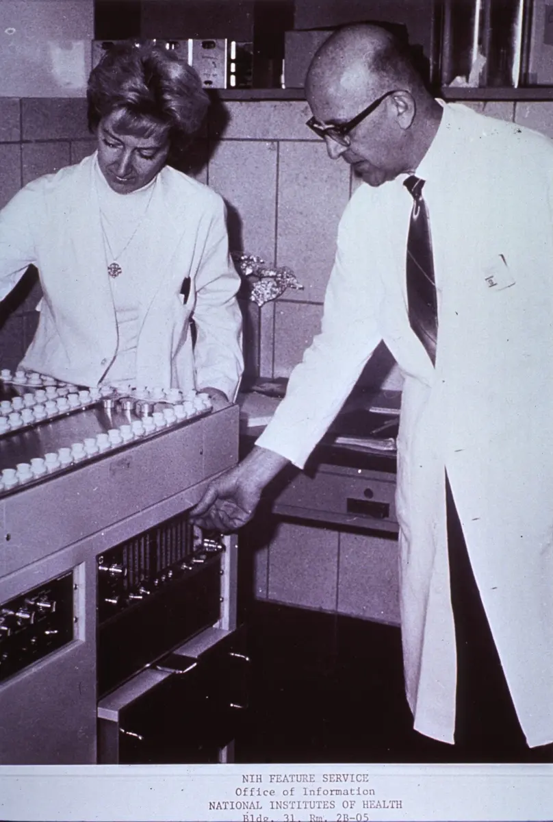 A woman and a man in white coats work at a large machine, pulling out a tray that holds rows of small tubes