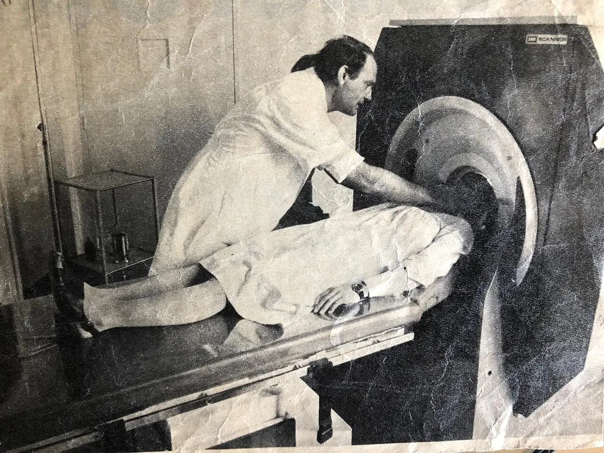 An old, creased black-and-white photo: a man in a white coat leans over a patient lying on a table, whose head is inside the round opening of a large dark machine