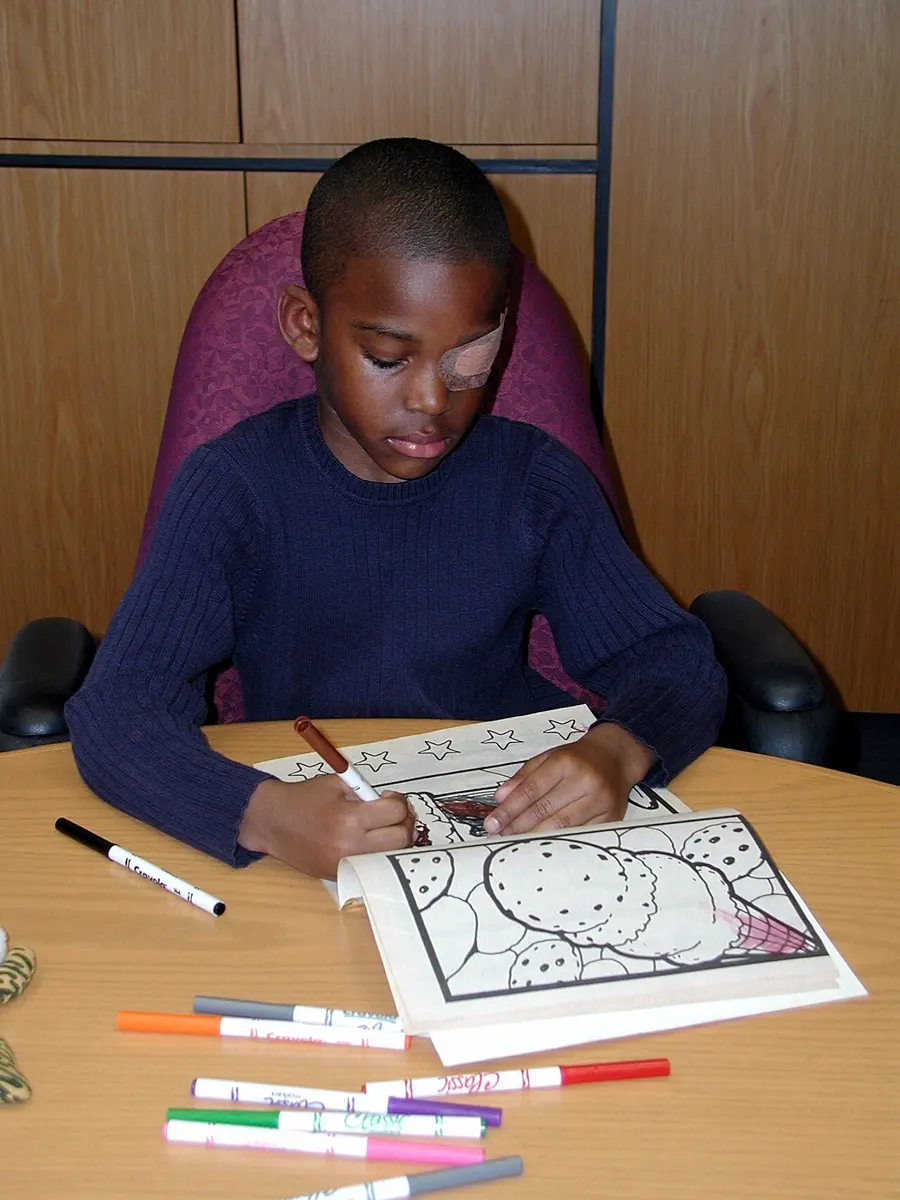 A boy sitting at a table, coloring a picture with felt pens, with a skin-colored patch over his left eye