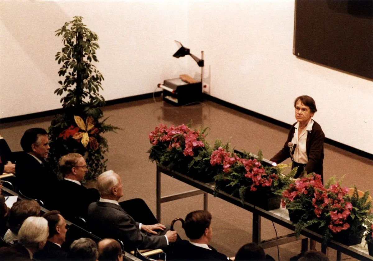 A woman with short gray hair in a dark jacket speaking at a desk with a microphone, in front of a raised hall full of people in suits; a long bank of pink flowers runs along the front