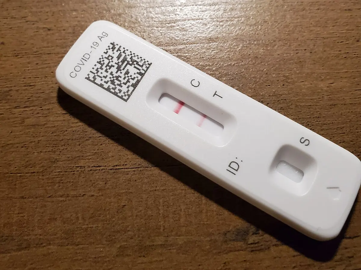 A white plastic test cassette labeled COVID-19 Ag lies on a wooden table; in its window are two red lines, beside the letters C and T