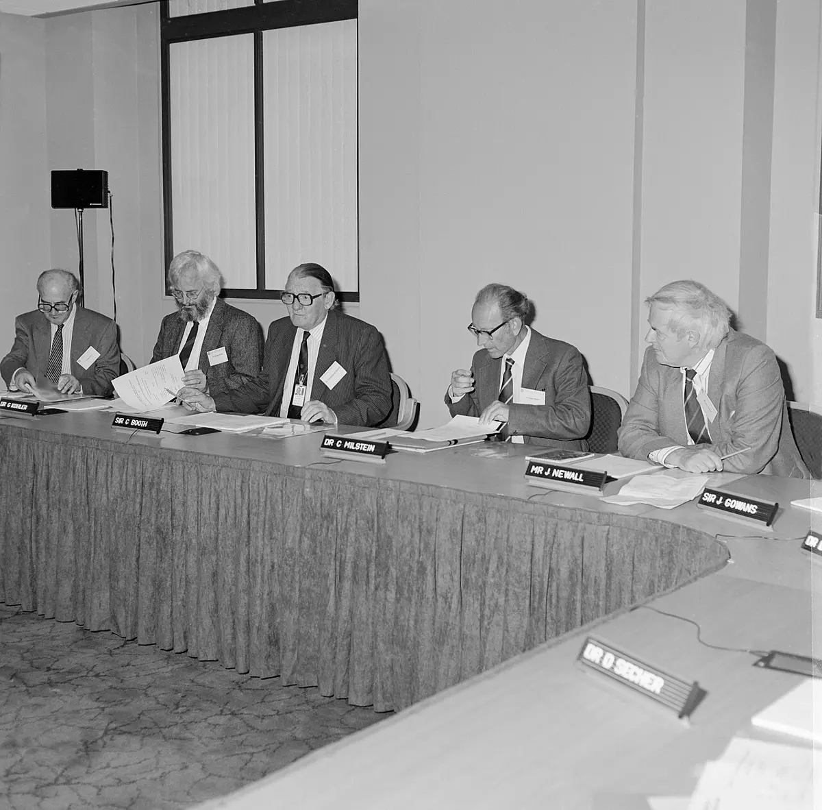 A black-and-white photo of five older men in suits seated at a long curved table with papers and name cards; the second from the left has a white beard and round glasses, the fourth wears dark-rimmed glasses
