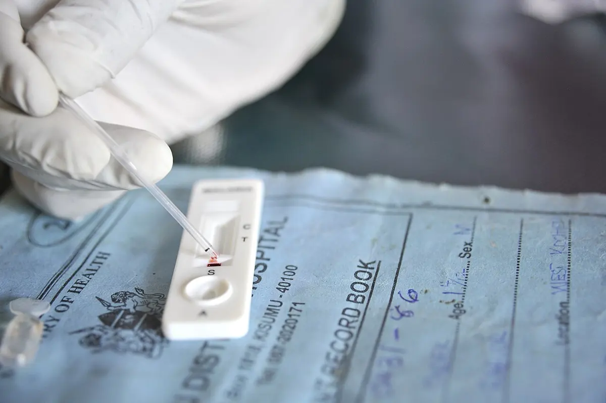 A gloved hand uses a thin dropper to put a drop of blood into the small well of a white plastic test strip marked C and T, which lies on a blue hospital record book