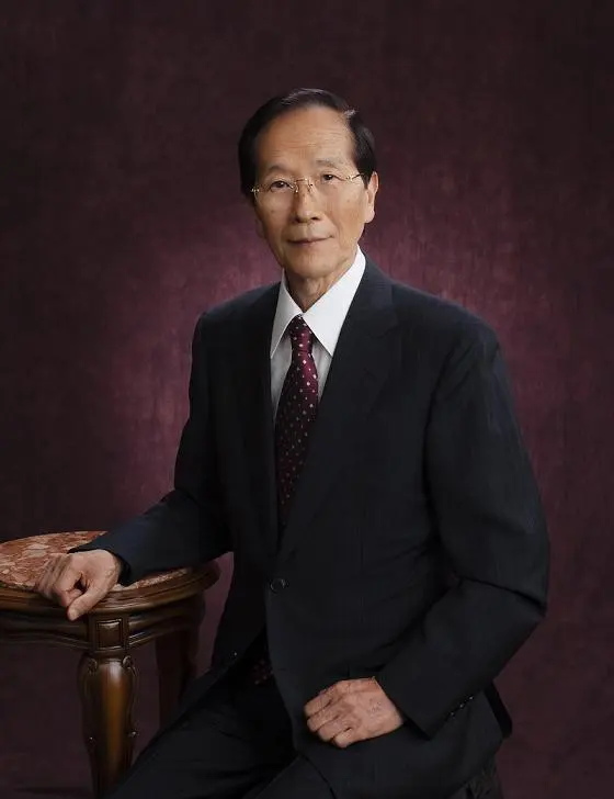 An elderly Japanese man with glasses, in a dark suit and tie, seated with one hand resting on a small wooden table