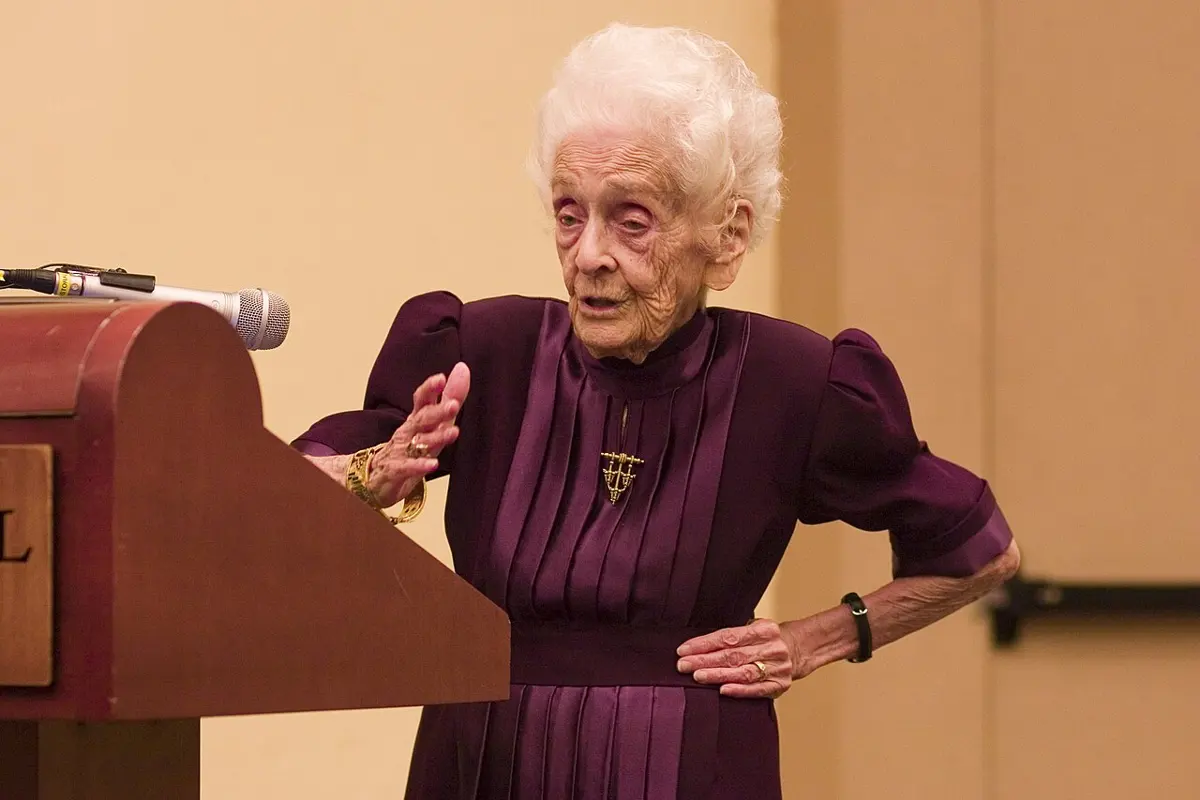 Rita Levi-Montalcini, aged 99, with white hair and a purple dress, speaking beside a lectern